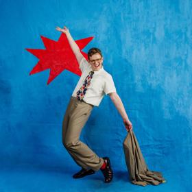 Kiwi comedian, Chris Parker, poses with his right arm raised, holding a brown suit jacket in his left hand in front of a blue background with a bright red spikey shape behind him.