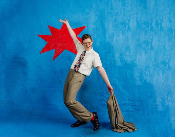 Kiwi comedian, Chris Parker, poses with his right arm raised, holding a brown suit jacket in his left hand in front of a blue background with a bright red spikey shape behind him.