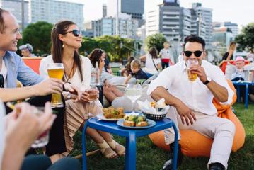 A group of four friends enjoy food and drinks on lawn furniture in a grassy field with city buildings in the distance.