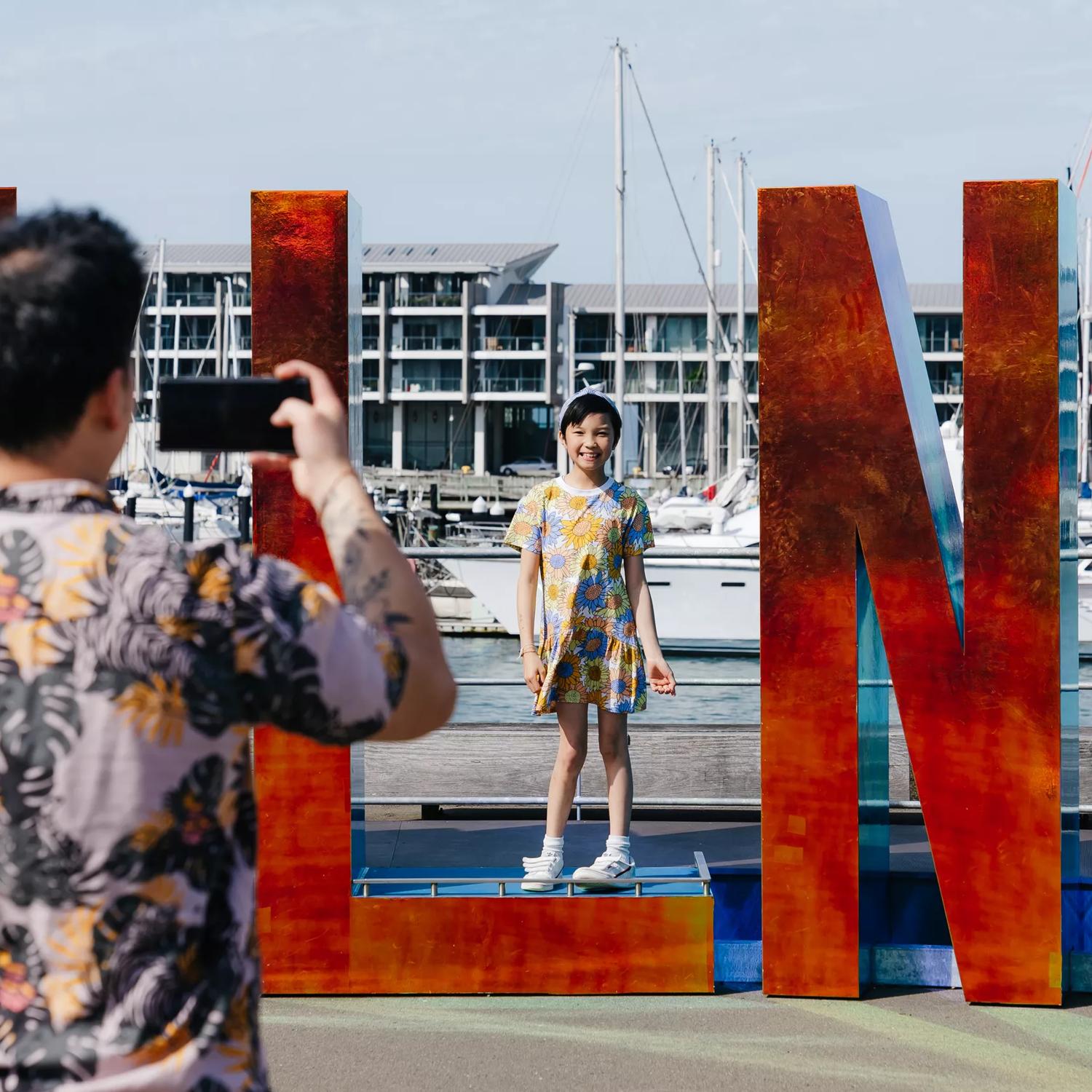 A parent taking of a photo of their child posing in the Well_ngton sign on the waterfront.