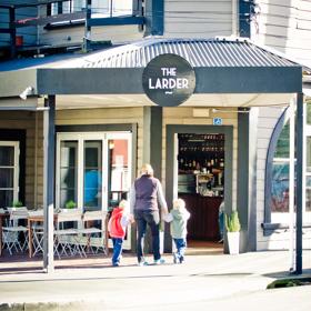 Exterior of The Larder café in Miramar. It is a grand old house with weatherboards and half-circle windows. Tables and chairs are outside and a parent and two children walk in the door.