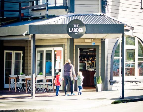 Exterior of The Larder café in Miramar. It is a grand old house with weatherboards and half-circle windows. Tables and chairs are outside and a parent and two children walk in the door.