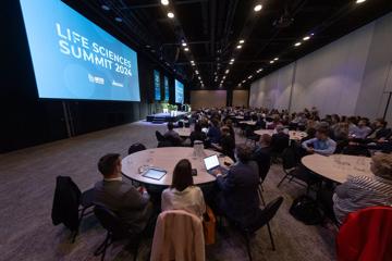 Inside Tākina Wellington Convention and Exhibition Centre the Life Sciences Summit 2024 is underway. Many people are seated in a large room set up with many round tables, a small stage and large projection screens.