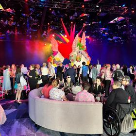 large conference room with colourful blue and pink lighting from the ceiling. in the foreground is a couch with people sat down and in the background is many people standing and talking