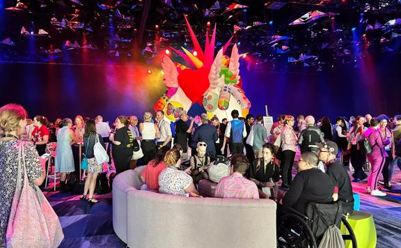 large conference room with colourful blue and pink lighting from the ceiling. in the foreground is a couch with people sat down and in the background is many people standing and talking