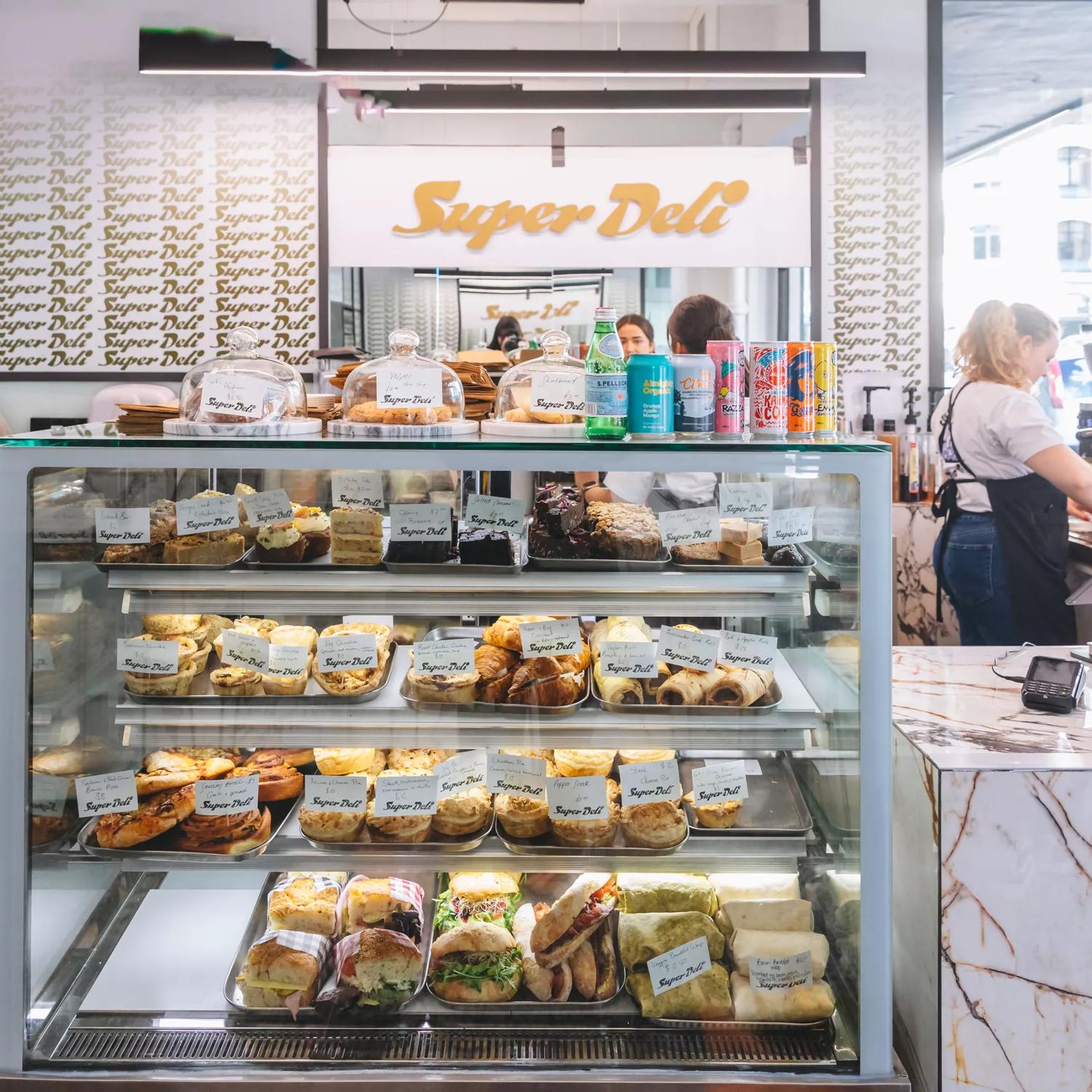 Inside Super Deli café, two workers are behind the counter and there is a glass case with sandwiched and baked goods on display.