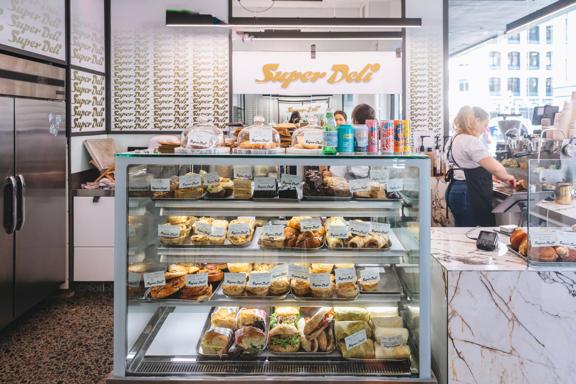 Inside Super Deli café, two workers are behind the counter and there is a glass case with sandwiched and baked goods on display.