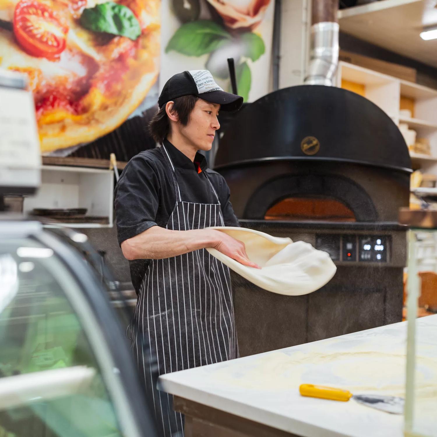 A person stands in the middle of a kitchen, stretching out pizza dough. A woodfire oven is in the background.