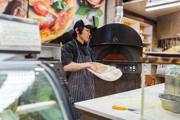 A person stands in the middle of a kitchen, stretching out pizza dough. A woodfire oven is in the background.