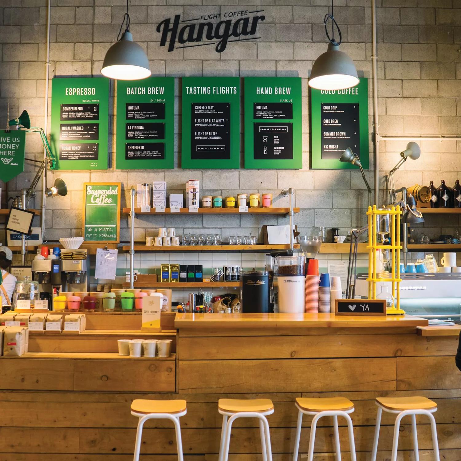 The front counter at The Hangar, made of wood with wooden stools and green menu boards on the wall, underneath their logo.