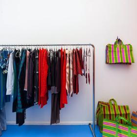 A rack of colourful clothing hangs against a wall at Sully’s. Colourful bags hang on a hook next to the rack.