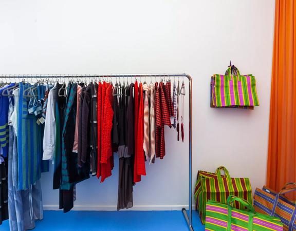 A rack of colourful clothing hangs against a wall at Sully’s. Colourful bags hang on a hook next to the rack.