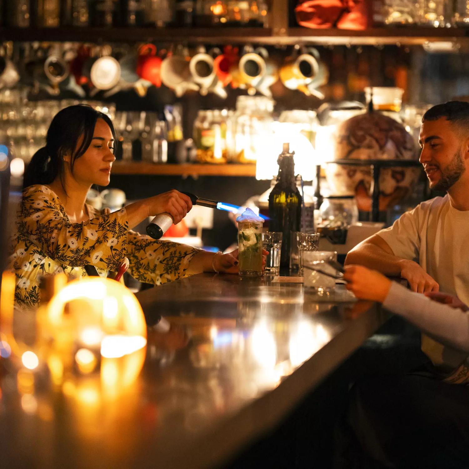 The interior of The Library, a cocktail bar on Courtney Place in Te Aro, Wellington. The bartender is using a flame to toast the garnish on a drink while two patrons are sitting at the bar.