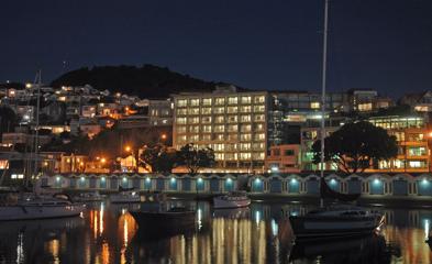 Wide shot over the Wellington harbour of the Copthorne Hotel on Oriental Pde, sailboats sit on the water in front of the colourful boat sheds and the hotel behind. Photo is taken at night time so the light from the building can be seen.