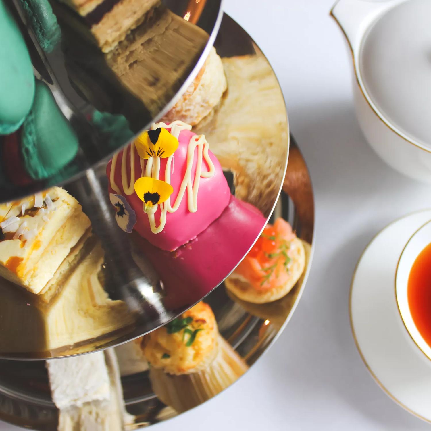 A silver cake stand with bite sized sweets sits on a table with a cup of tea.