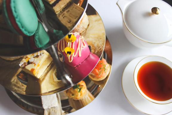 A silver cake stand with bite sized sweets sits on a table with a cup of tea.