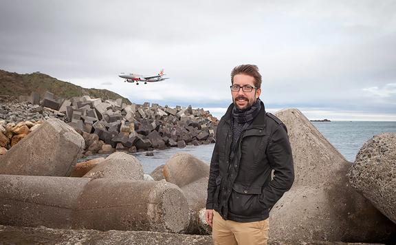 Episodic director of TV show ‘Thunderbirds Are Go’ stands in front of rocks and the ocean in Lyall Bay as a plane comes into land.