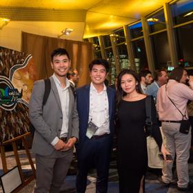 Three delegates in semi-formal wear stand together and smile for a picture being a large painting on a easle at the ICMS Australasia dinner.