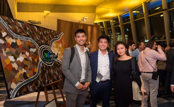 Three delegates in semi-formal wear stand together and smile for a picture being a large painting on a easle at the ICMS Australasia dinner.