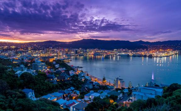 The view of Wellington harbour from the look out point on Mount Victoria at dusk. The sky is purple with a hint of orange glow from the sun in the distance and the city street lights are illuminated. 