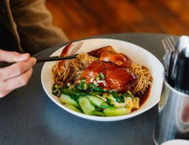 A diner uses chopsticks to lift a slice of roasted duck from a bowl of noodles topped with bok choy and scallions, served on a café table beside a container of cutlery.