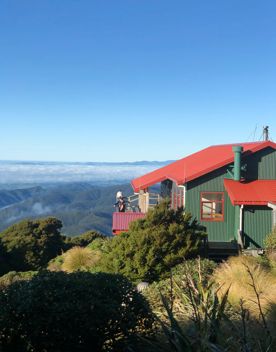 Powell Hut in Tararua Forest Park in the Wairarapa region. It’s a small house with dark green siding, and a red roof with two people standing on the exterior wooden deck.
