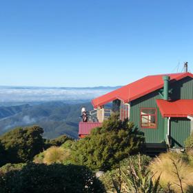 Powell Hut in Tararua Forest Park in the Wairarapa region. It’s a small house with dark green siding, and a red roof with two people standing on the exterior wooden deck.