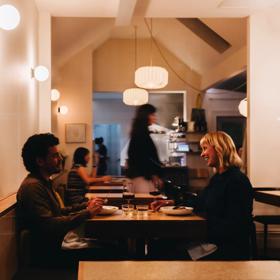 Two friends enjoy a meal at Rita. The small restaurant has dim amber lighting and staff working in the background.