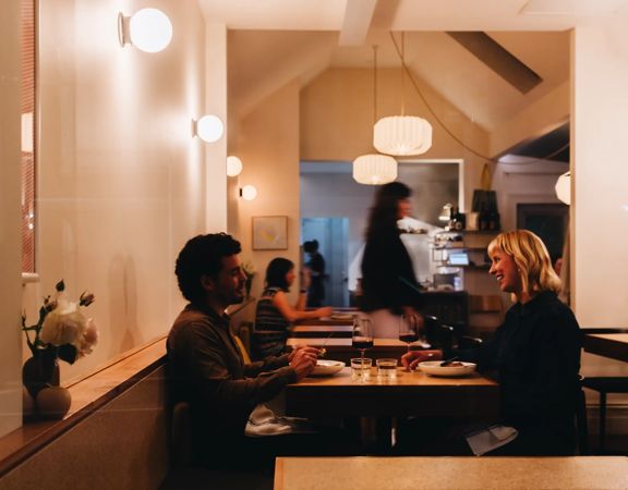 Two friends enjoy a meal at Rita. The small restaurant has dim amber lighting and staff working in the background.