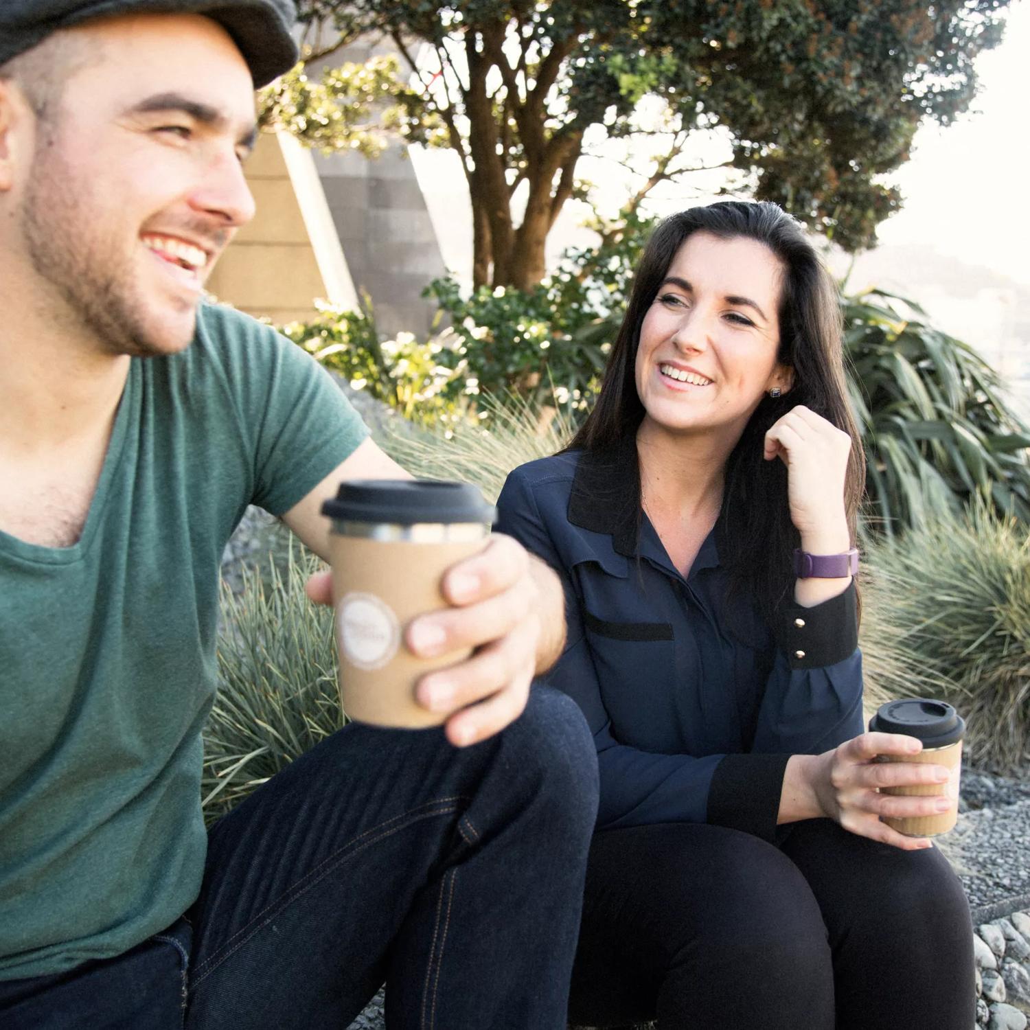 Two people sit on the Wellington waterfront holding reusable coffee cups from Again Again.