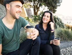 Two people sit on the Wellington waterfront holding reusable coffee cups from Again Again.