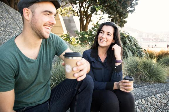 Two people sit on the Wellington waterfront holding reusable coffee cups from Again Again.