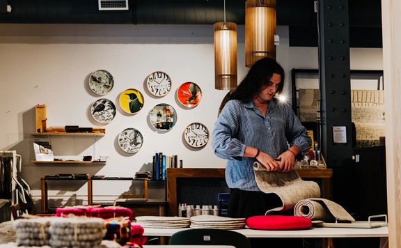 Person arranging handcrafted felt products on a table, with colourful wall plates and shelving in the background.