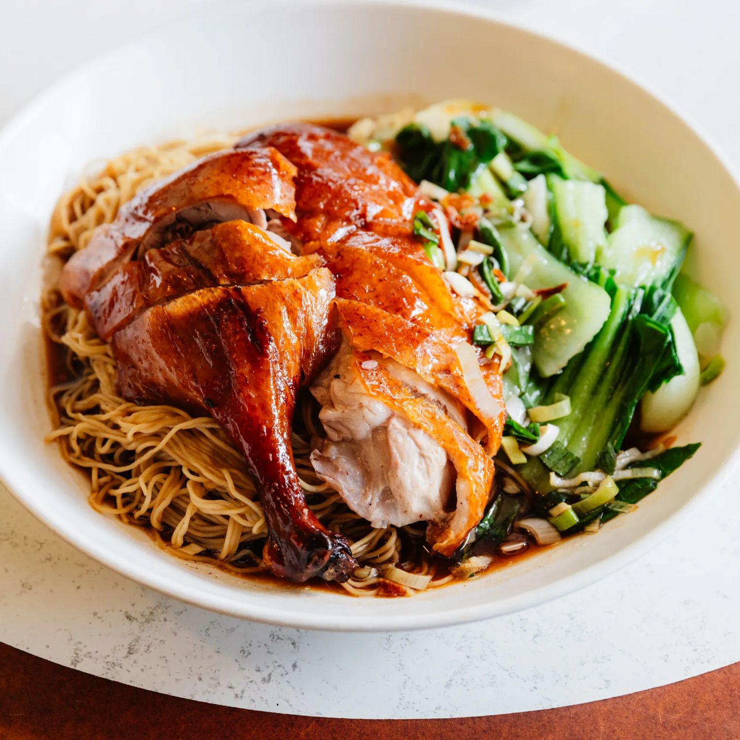 A bowl of roasted duck served over thin egg noodles with bok choy and scallions, topped with sauce and presented on a white tabletop.