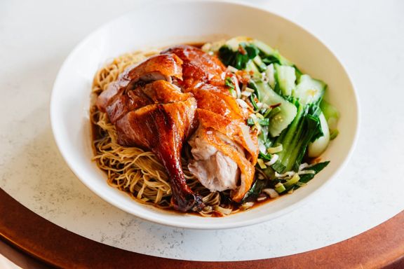 A bowl of roasted duck served over thin egg noodles with bok choy and scallions, topped with sauce and presented on a white tabletop.