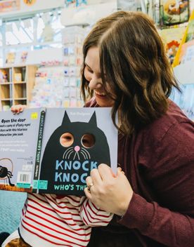 An adult holds a baby in a bookstore while showing a children’s book titled “Knock Knock Who’s There?”, with shelves of colourful books in the background.