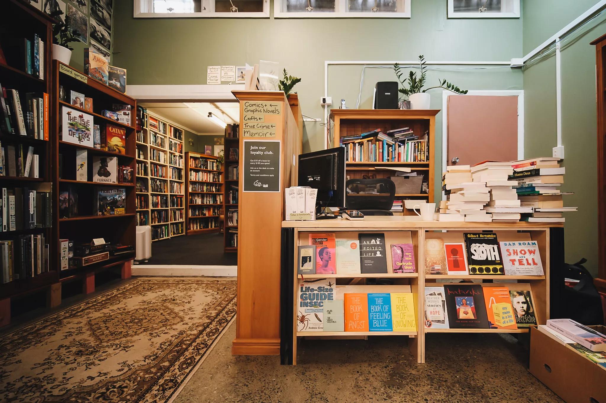The check-out counter at Book Hound, a bookstore in Newtown, Wellington. The small space has pale green walls, a Persian rug, and wooden shelves filled with books. 
