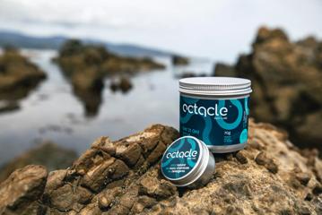 Two tins of Octacle wetsuit lube, one large and one small, on a rock with water and rocks in the background.