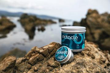 Two tins of Octacle wetsuit lube, one large and one small, on a rock with water and rocks in the background.