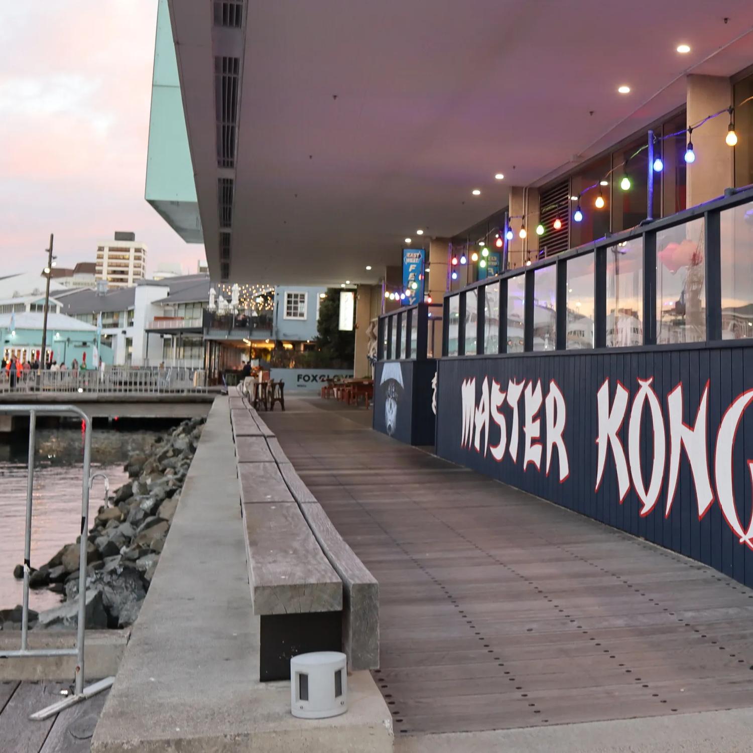 The exterior of Master Kong, a bar & restaurant on Wellington's waterfront during golden hour. There are colourful string lights and paper lanterns suspended above the patio. 