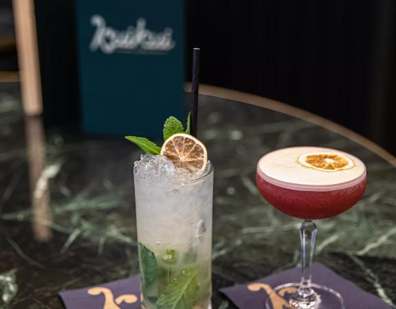 2 cocktails are placed on a green marble table, one drink is white with green garnish and the other is red with a lemon garnish.