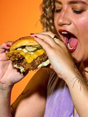 Person biting into a cheeseburger topped with pickles against an orange background.