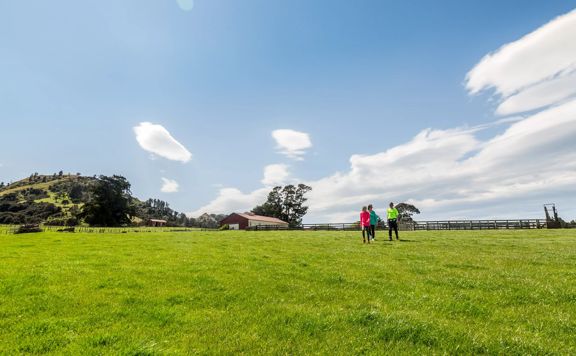Three people in neon shirts, walking across an expansive grass field. There is a red farm building and wooden farm fencing in the background.