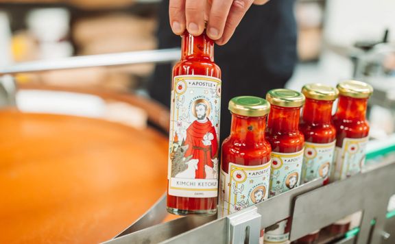 A hand holds a bottle of Apostle Hot Sauce Kimchi Ketchup at their headquarters in Paraparaumu, and there are four other bottles in a row on the metal packaging track.