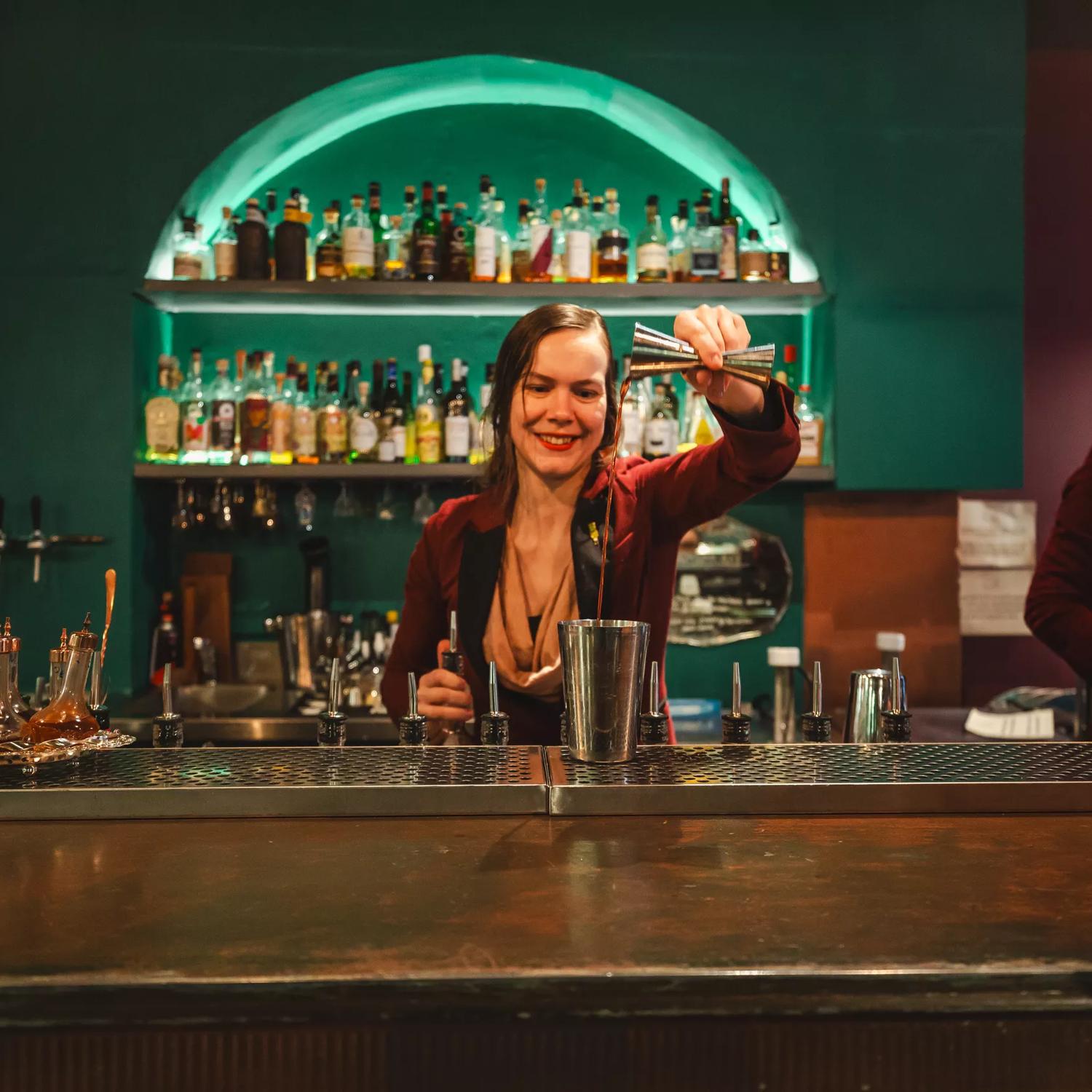 A bartender pours liquor into a cocktail shaker. Alcohol bottles line the illuminated shelves behind them.