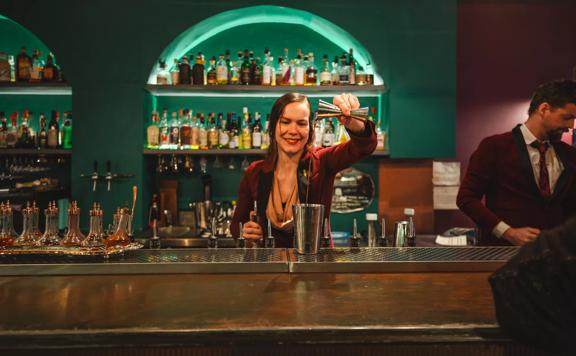 A bartender pours liquor into a cocktail shaker. Alcohol bottles line the illuminated shelves behind them.