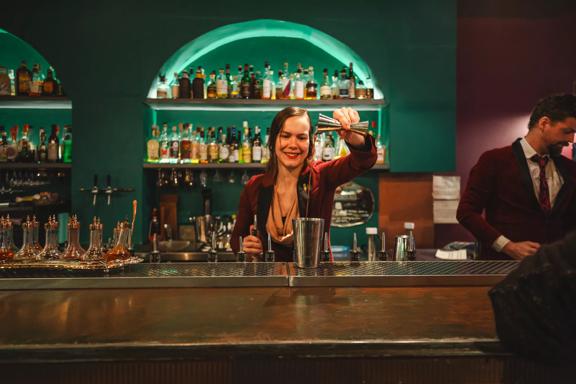 A bartender pours liquor into a cocktail shaker. Alcohol bottles line the illuminated shelves behind them.