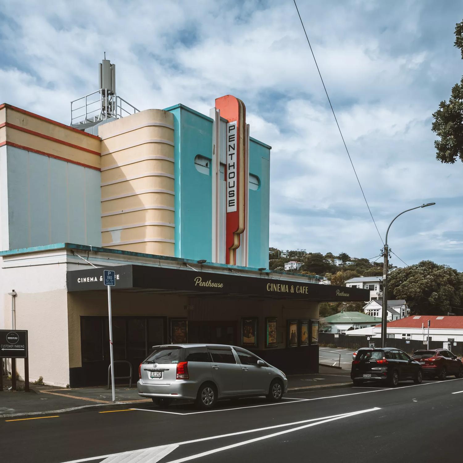 Penthouse Cinema & Cafe shown from the street looking towards the front of the building. The building is art deco style, with aqua, peach and red architectural detailing.