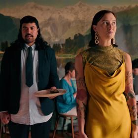 2 people standing in a restaurant, one of them holds a plate of food, wearing a suit, and the other wears all yellow.