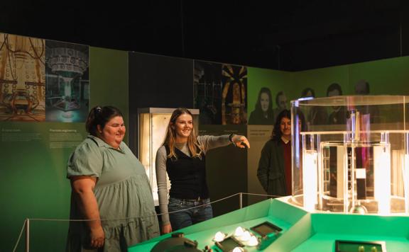 Three people smiling as they stand inside the Tardis control room. The room has green walls, and the person in the middle is pointing at a glass box on the control panel.